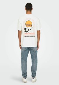 Camiseta blanca de algodón con un diseño gráfico en la parte posterior que presenta una ilustración de una hamburguesa y personajes de dibujos animados en acentos negros y rosas.