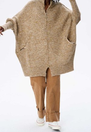 Personne portant un pull en maille beige surdimensionné zippé avec des poches, un pantalon marron à jambes larges et des baskets blanches marchant vers l'avant.