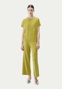 Limegrön stickad topp och vida byxor med ribbad textur. Metallic klackskor kompletterar outfiten. Modellen står emot en ljus bakgrund.