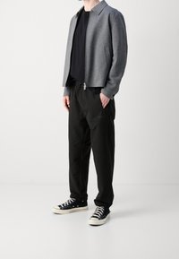Persoon in een grijze ritsjas, zwarte t-shirt, zwarte broek met trekkoord en zwarte hoge sneakers met witte zolen en veters.