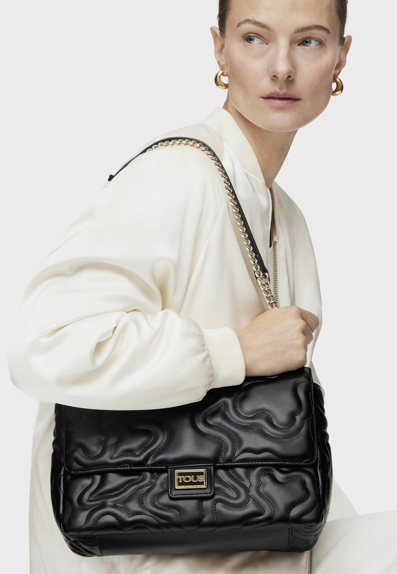 Borsa in pelle nera con design trapuntato, hardware dorato e tracolla a catena. Texture liscia, ampio scomparto principale, placca con logo del brand in evidenza.