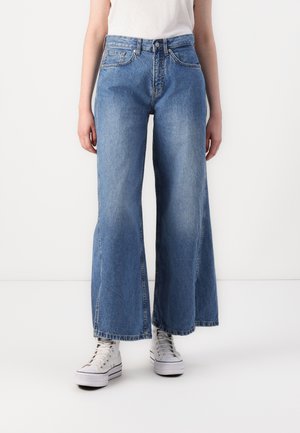 Jean en denim bleu à jambes larges avec une taille haute, présentant des poches avant et un délavé. Associés à des baskets montantes blanches.
