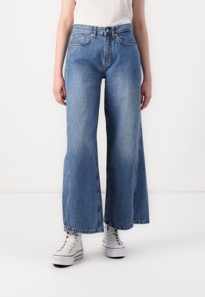Jean en denim bleu à jambes larges avec une taille haute, présentant des poches avant et un délavé. Associés à des baskets montantes blanches.