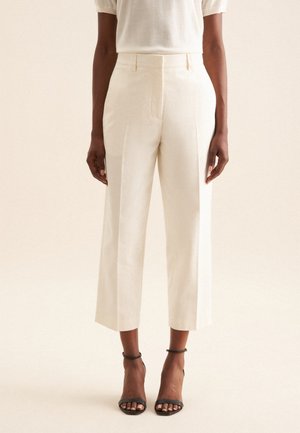Pantalons taille haute de couleur crème, ajustés, avec une jambe raccourcie, présentant des plis et des poches latérales, associés à des sandales noires à bride cheville.
