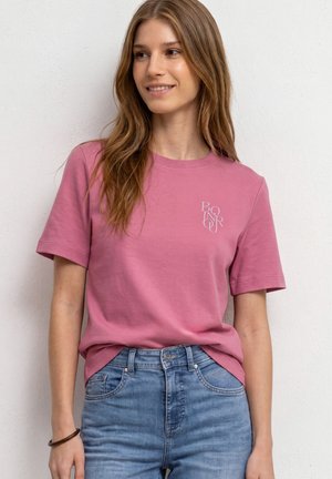 Ung kvinde med langt brunt hår iført en pink t-shirt med teksten "BONJOUR" og blå jeans, smilende let mens hun kigger til siden.
