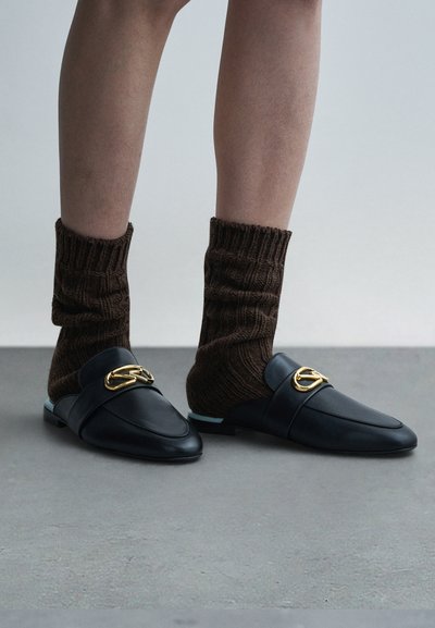 Mustad nahast mokassiinid kuldse dekoratiivse detailiga, pruunide soonikkoeliste sokkidega, hallil taustal.