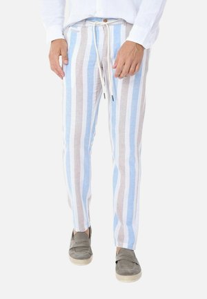 Uomo che indossa pantaloni beige, bianchi e blu a righe verticali con coulisse e mocassini beige, in piedi su sfondo bianco.