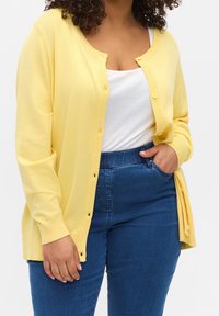 Donna con un cardigan giallo abbottonato sopra una canotta bianca a coste, che indossa jeans blu a vita alta, con una mano in tasca.