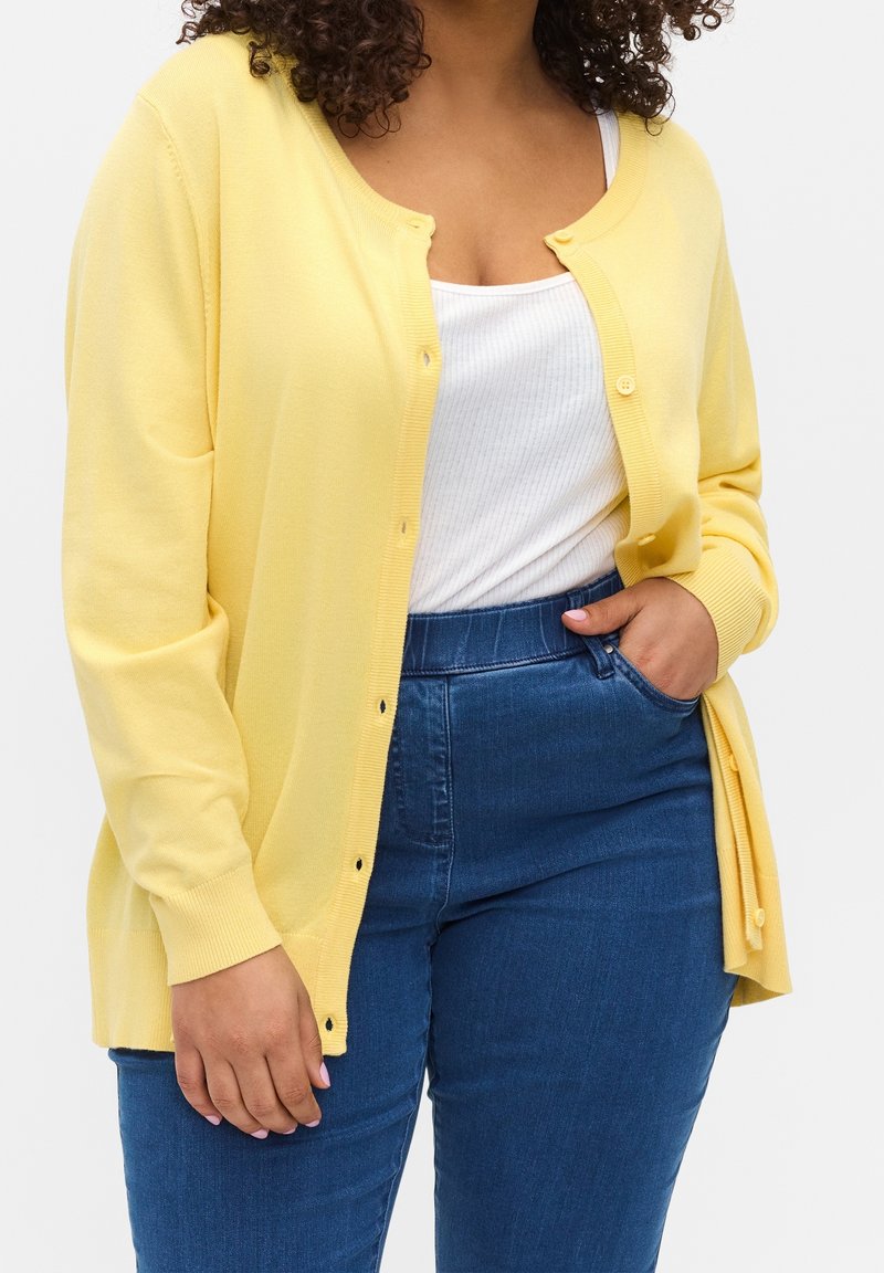 Donna con un cardigan giallo abbottonato sopra una canotta bianca a coste, che indossa jeans blu a vita alta, con una mano in tasca.