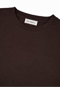 Tummanruskea puuvillapaita, jossa on pyöreä pääntie ja lyhyet hihat. Edessä on hillitty brodeerattu logo "Oh April".