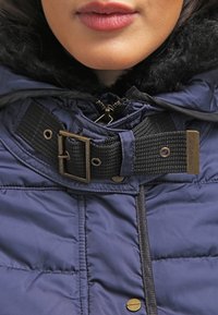 Chaquetón de marino con un cinturón texturizado negro y hebilla de latón. Presenta una capucha y un cierre de cremallera con broches dorados en la parte delantera.