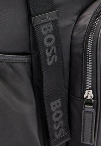 Black nylon backpack featuring a woven strap labeled "BOSS," silver zippers, and a smooth texture with a side pocket detail.