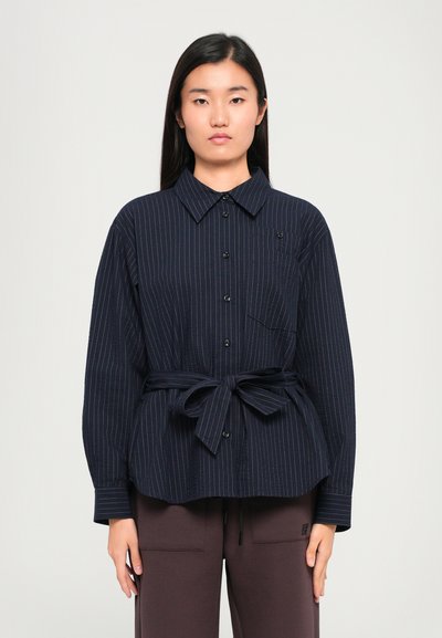Chemise boutonnée bleu marine à rayures avec col, manches longues et taille nouée. Présente une poche poitrine et des boutons noirs.