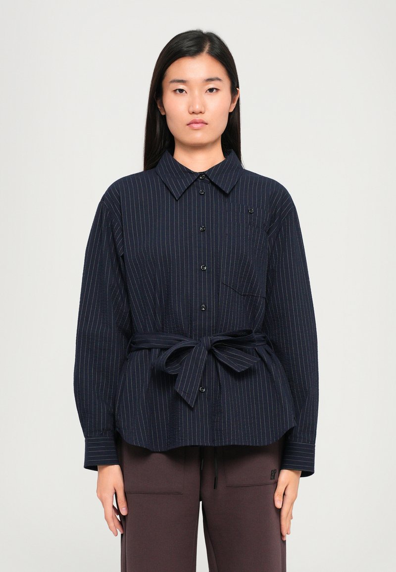 Navy pinstriped button-up shirt with a collar, long sleeves, and a tied waist. Features a chest pocket and black buttons.
