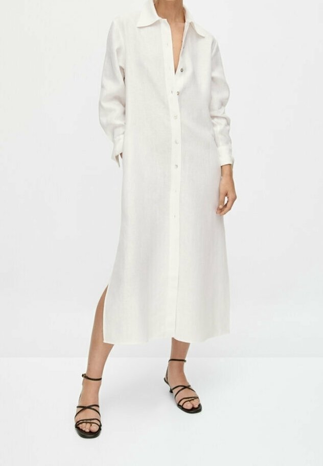 Femme portant une longue robe chemise blanche à boutons avec des fentes latérales, associée à des sandales plates noires à fines lanières.