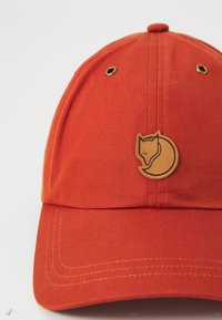Rustfarvet baseballkasket med en tan cirkulær lap, der viser et abstrakt rævehoved på frontpanelet.