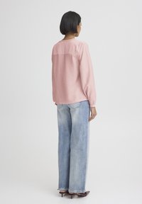 Růžová halenka s dlouhým rukávem a texturovanými zády, ve spojení s světle modrými džínami se širokými nohavicemi a tmavými podpatky, viděná zezadu.