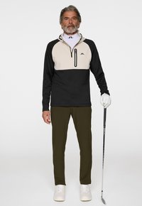 Pull-over bicolore pour homme en crème et noir avec une demi-fermeture éclair, associé à un pantalon vert olive et des baskets blanches. Tenant un club de golf.