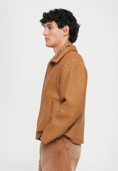 Chaqueta marrón con canalé y textura suave, con cuello y bolsillos laterales, llevada sobre unos vaqueros claros.