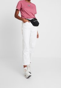 Růžové tričko s krátkým rukávem s logem, bílé džíny rovného střihu a černá ledvinka; bílé tenisky s růžovými akcenty a texturovanou podrážkou.