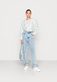 Blusa azul clara de padrão floral com mangas bufantes, combinada com jeans de lavagem clara de cintura alta. Modelo segura uma bolsa azul clara e usa ténis brancos.