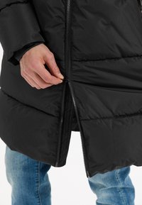 Sort puffjakke med lynlås, der har et quiltet design og en høj krave. Håndjustering af lynlåsen i kanten.