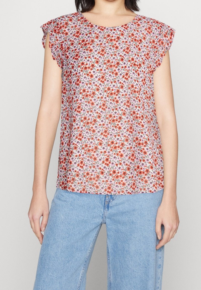 Femme portant un haut blanc sans manches avec un imprimé floral rouge et orange et un jean taille haute bleu clair sur un fond uni.