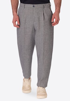 Uomo con pantaloni grigi plissettati e sneakers beige in piedi su sfondo bianco.
