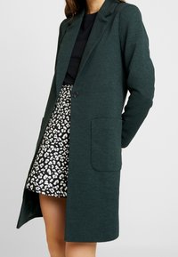 Zelený texturovaný kabát s klopovým límcem, jedním knoflíkem a bočními kapsami, v kombinaci s sukní se vzorem černobílé leopardí kůže.