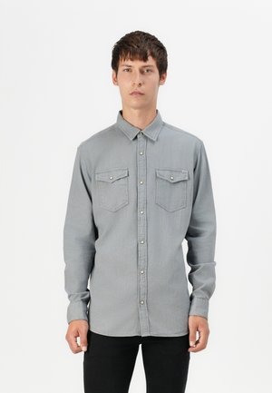 Chemise en denim gris avec un col boutonné, deux poches sur la poitrine et des boutons argentés. Elle présente des manches longues et une coupe ajustée.