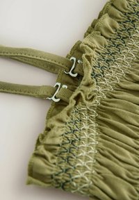 Kostkovaná tkanina olivové barvy se smocking texturou, má nastavitelné ramínka a jemné modré akcenty, které dotváří design s volánky.