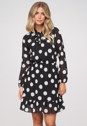 Femme blonde portant une robe noire à manches longues avec de grands pois blancs, une ceinture à la taille, un ourlet froncé et un nœud au col.