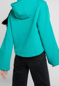 Haut court turquoise avec capuche et manches larges, fabriqué en tissu doux. Associé à un pantalon noir taille haute avec des détails texturés.