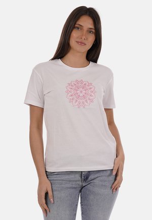 Donna con capelli lunghi e scuri che indossa una t-shirt bianca a maniche corte con un complesso disegno floreale mandala rosa, abbinata a jeans azzurri.