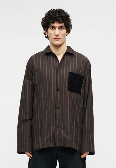Νεαρός άνδρας με κατσαρά μαλλιά, φοράει ένα φαρδύ καφέ πουκάμισο με ρίγες και μια μεγάλη υφασμάτινη μαύρη τσέπη στο αριστερό στήθος.