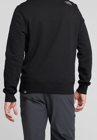 Černá mikina s kulatým výstřihem, pružnými manžetami a lemem. Na horní části ramene se nachází malé logo, vyrobené z měkkého texturovaného materiálu.