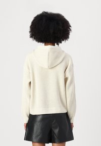 Felpa con cappuccio realizzata in filato crema texturizzato, caratterizzata da vestibilità ampia, spalle scese e orlo a coste. Indossata con pantaloncini neri in ecopelle.