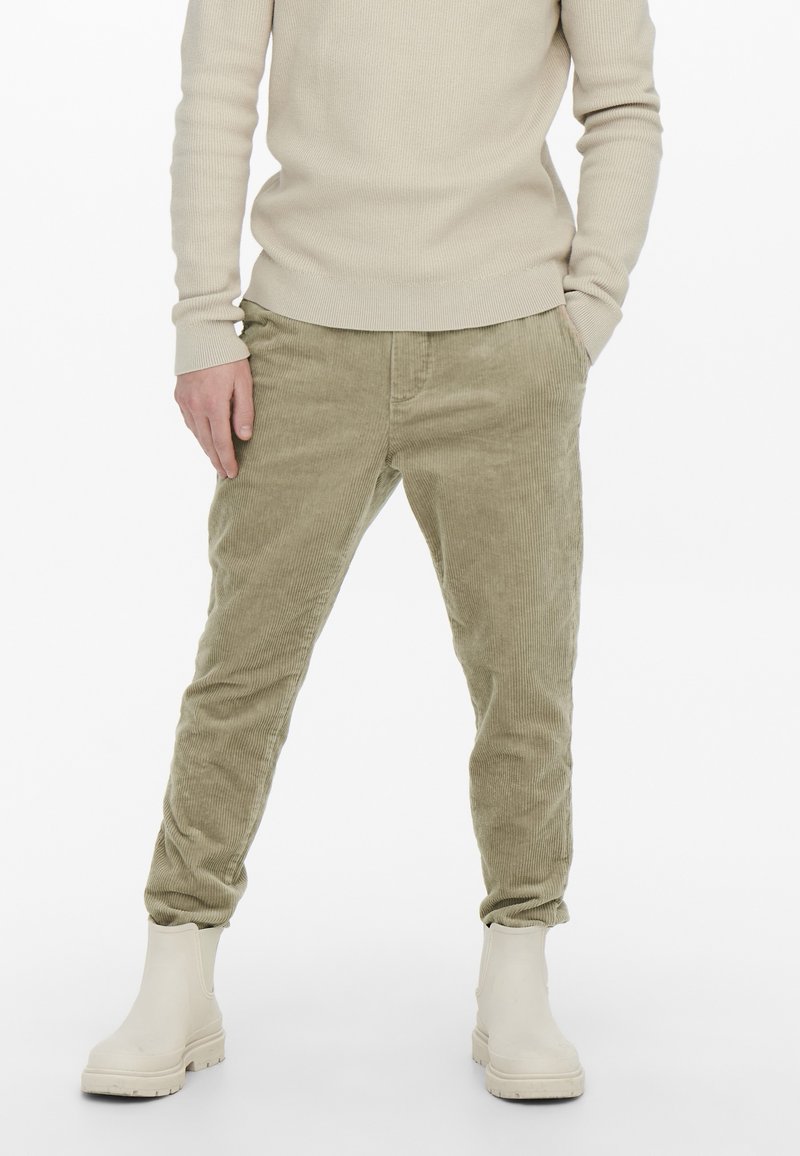 Personne portant un haut beige côtelé à manches longues, un pantalon en velours côtelé marron clair et des bottes épaisses écrues, les mains partiellement dans les poches.