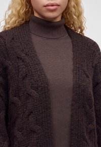 Personne blonde portant un cardigan en maille torsadée marron foncé sur un pull à col roulé marron assorti, montré de la poitrine aux lèvres.
