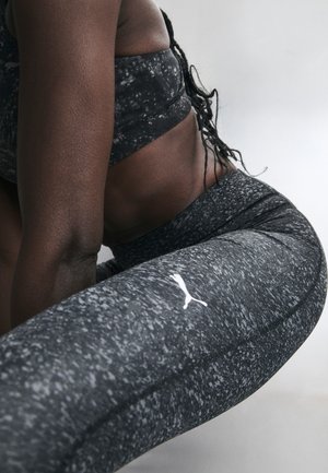 Gros plan d’une personne en tenue de sport noire à motifs qui s’étire, montrant son torse et sa jambe avec un logo Puma blanc sur les leggings.