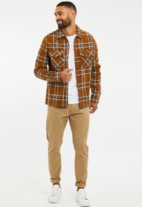 Brown and plaid flannel shirt with two chest pockets, paired with beige chinos and white sneakers. The shirt has a button-up style.