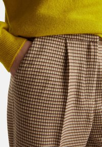 Bruine geruite broek met crème houndstooth patroon, voorzien van zijzakken en plooien. Gecombineerd met een felgele gebreide trui.