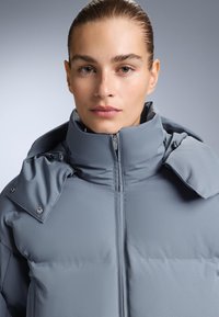 Siva podstavljena jakna s visokim ovratnikom, zatvaranjem na patentni zatvarač i detaljima na kopčanje. Glatka tkanina s blago sjajnim završetkom.