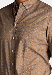 Bruine button-up blouse met een klein, herhalend patroon van blauwe en oranje ovalen. Bevat een borstzak en witte knopen.