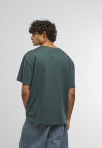 Tmavě zelené bavlněné tričko, volný střih, kulatý výstřih, krátké rukávy, jednoduchý design, kombinováno s modrými džíny, viditelné švy.