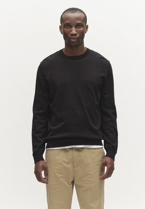 Man staat naar voren gericht en draagt een zwarte trui met lange mouwen over een wit overhemd en beige broek tegen een effen witte achtergrond.