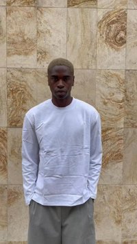 Giovane uomo con capelli biondi corti, che indossa una camicia bianca a maniche lunghe e pantaloni grigi, in piedi di fronte a una parete di marmo beige.
