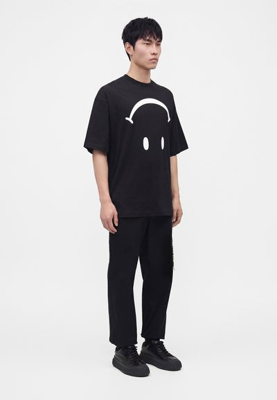 Jeune homme debout portant un T-shirt noir oversize avec un graphisme blanc de casque et d'œil, pantalon noir et baskets noires sur fond blanc.