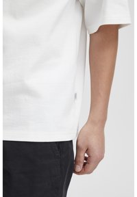 Hvid bomulds t-shirt med korte ærmer. Sømmene har et lille brandmærke. Personen bærer sorte bukser. Glat overflade generelt.