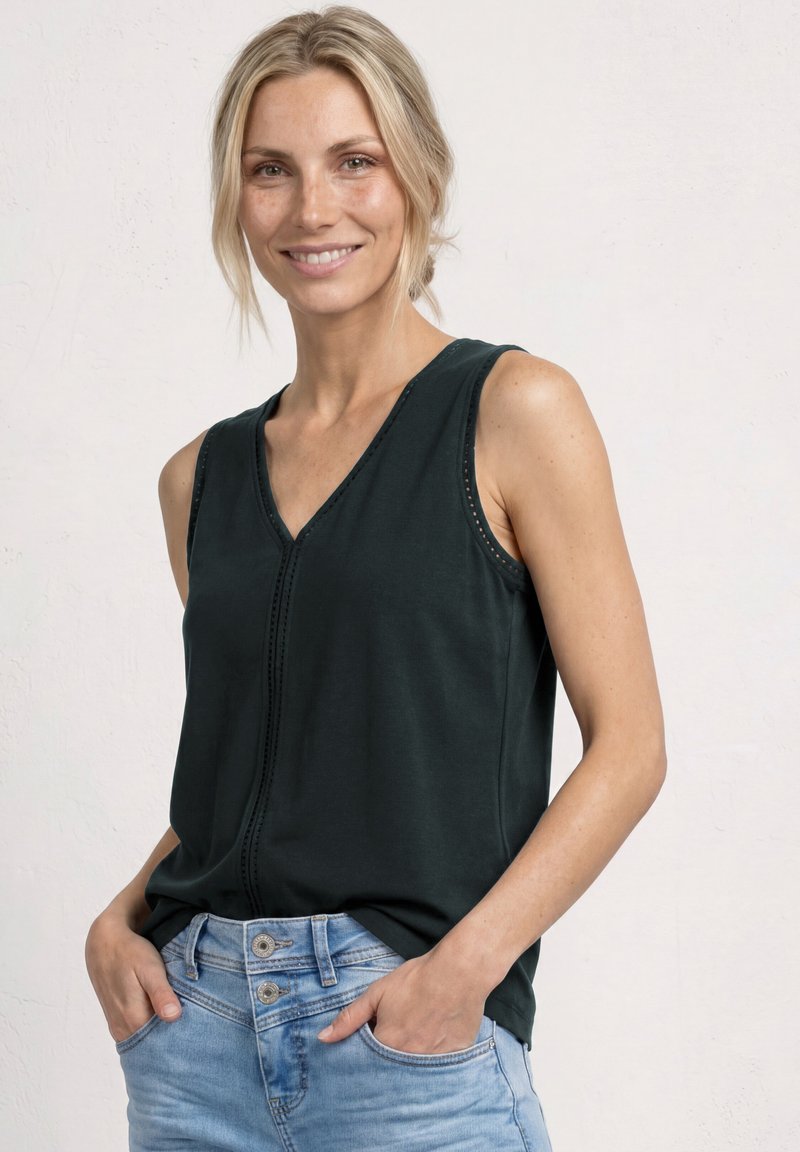 Femme souriante aux cheveux blonds portant un haut noir sans manches et un jean bleu clair, les mains dans les poches, debout devant un fond uni.